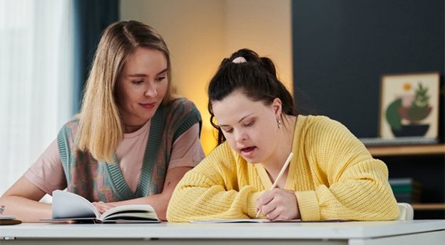 A person with Down syndrome writing while someone else looks on.