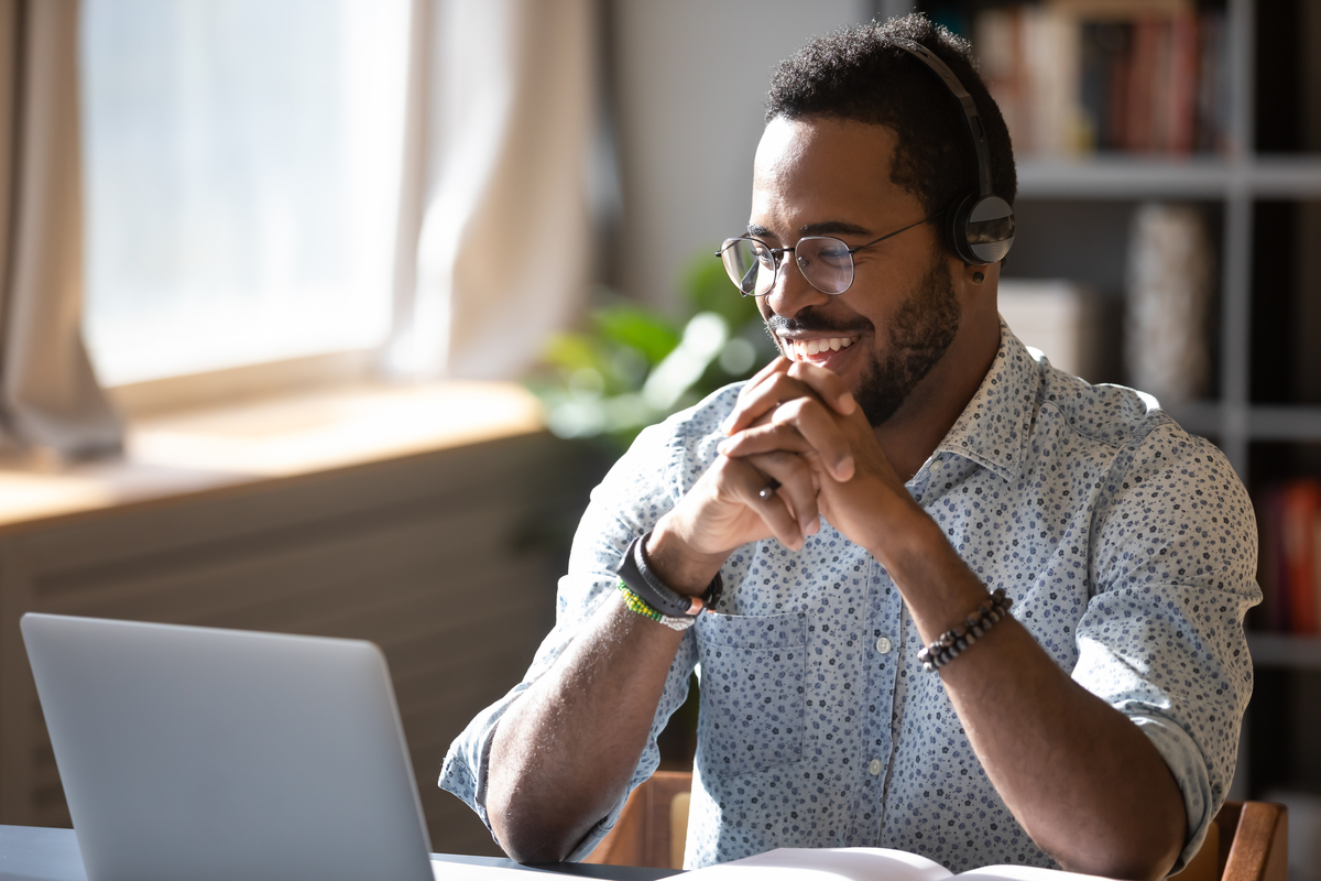 Happy millennial African American man in glasses wearing headphones, enjoying watching educational webinar on laptop.