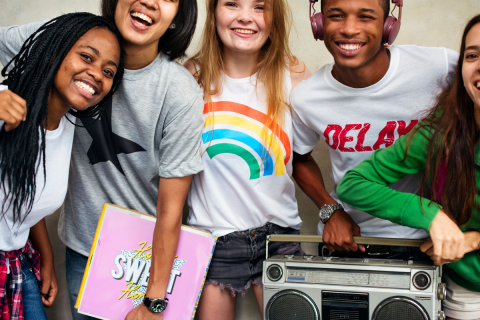 Group of 5 diverse teens smiling. One holds a radio, another holds a binder.