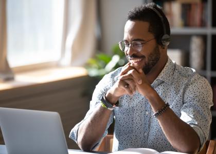 Happy millennial African American man in glasses wearing headphones, enjoying watching educational webinar on laptop.