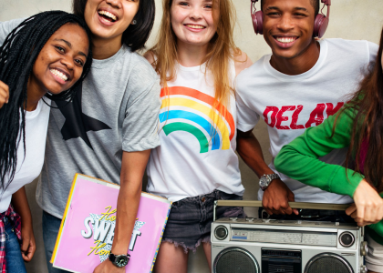 Group of 5 diverse teens smiling. One holds a radio, another holds a binder.