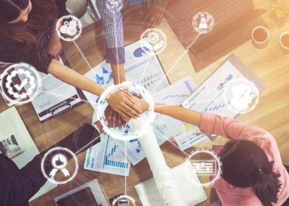 Image taken from above of 5 peoples hands joined in the center over a table stren with office supplies and computers. An overlay shows circle around the hands with images of gears, charts, check mark, person connected as spokes. 