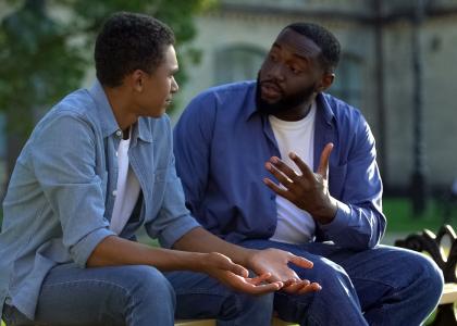 Man sitting on a park bench talking to teenage boy.