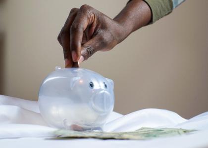 Image of a hand putting money into a clear piggy bank.