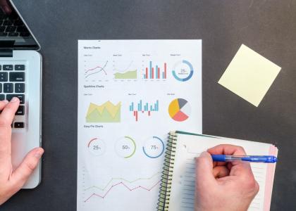 Image of a hand taking notes in a notebook that rests upon a piece of paper with various graphs