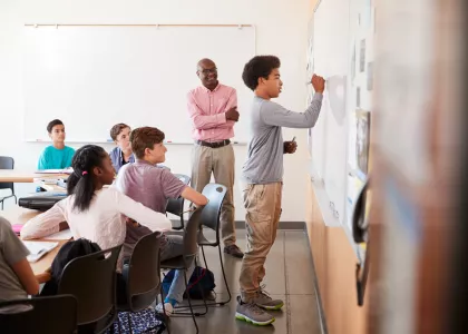 A male teacher in a classroom of adolescents. One teen is working a problem at the whiteboard as other students and the teacher observe. 
