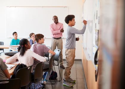 A male teacher in a classroom of adolescents. One teen is working a problem at the whiteboard as other students and the teacher observe. 