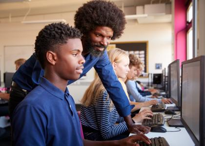 Male teacher works at computer station with teen boy.