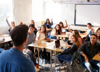 Happy students in a classroom facing an instructor. They are raising their hands to respond. 