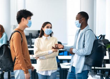 Three young adults wearing face masks standing a classroom talking.