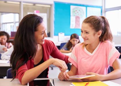 Smiling female teacher sits at a desk in a classroom beside a smiling teen girl